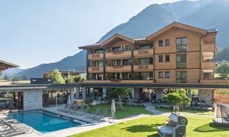 A modern building in alpine style with several balconies. In the foreground is a pool and a green lawn area.