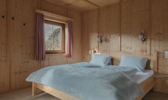 A cozy bedroom made of wood with a large bed and blue blankets. The windows with curtains let in soft light.