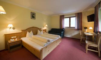 A cozy room with two beds, a sofa, and a table. The walls are painted yellow, and large windows provide plenty of daylight.