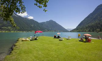 A green meadow by the shore of a clear lake, surrounded by mountains. Sun loungers and umbrellas invite you to relax.