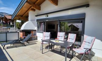 A cozy terrace with a table and chairs, ideal for relaxing. In the background, there is a grill, and the surroundings are characterized by wooden details and single-family homes.