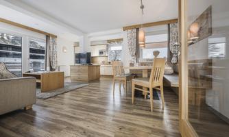 A modern living room with wooden flooring and large windows. The furnishings include a dining area with a wooden table and chairs, as well as a cozy seating area.