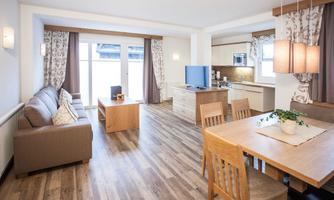 Bright and modern living room with a cozy couch and a dining table. The open kitchen is well-equipped and the large windows provide plenty of natural light.