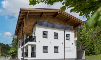 A modern building in alpine style with a visible wooden roof. It has several windows and balconies as well as a quiet environment with trees.