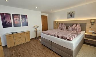 A modern bedroom with a large bed and red-and-white checked pillows. Pictures of forests hang on the wall, and the room has wooden flooring.