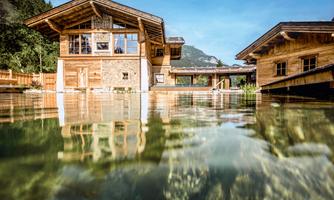 A modern wooden house by the water with large windows and a beautiful surroundings. In the background, forested mountains can be seen.