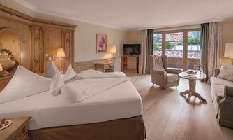 A cozy hotel room with a double bed and traditional wooden furniture. The room has large windows overlooking nature and is lovingly decorated.