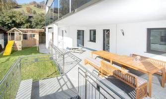 A modern garden area with a wooden terrace and a large dining table. In the background, there is a playhouse with a slide visible.