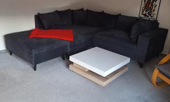 A modern living room corner with a large, dark gray sofa and a white coffee table. Some pictures hang on the wall, and the floor is covered with gray carpet.