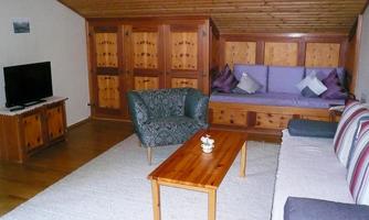 A cozy living room with wood paneling and a large window. There is a sofa, an armchair, and a wooden table.