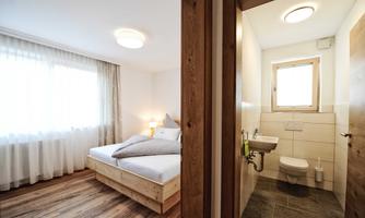 A cozy bedroom with a large bed and a window that lets in daylight. Adjacent is a modern bathroom with sanitary facilities.