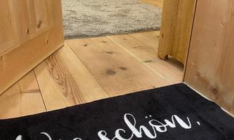 An inviting entrance with a black carpet that says: "How nice to have you here." In the background, there is a wooden chair and a cozy rug.