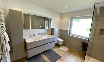 A modern bathroom with a white sink and a wide mirror. The room features wooden flooring and plenty of daylight through a window.