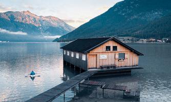 A wooden house on stilts in the water, surrounded by mountains. A boat is nearby the dock.