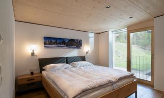 A modern bedroom with a large bed and wooden ceiling. Large windows offer a view of the greenery.
