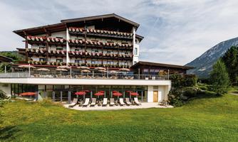 A modern hotel surrounded by green grass and trees. On the terrace, there are loungers with sunny umbrellas.