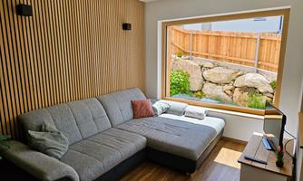 A modern living room with a gray couch and a television. Through a large window, you can see a green garden with stones and a wooden fence.