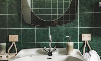 A modern bathroom with an elegant sink and a round mirror. The walls are decorated with green tiles.