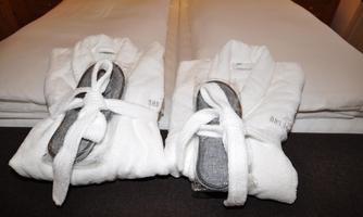 Two well-groomed bathrobes and slippers are neatly placed on a bed. In the background, white bed linen and a cozy atmosphere can be seen.