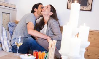 A couple is sitting comfortably together and laughing. Snacks and a glass of wine can be seen on the table.
