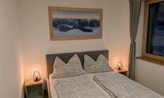 A cozy bedroom with a large bed and modern pillows. Decorative pictures hang on the walls, and soft light creates a relaxed atmosphere.