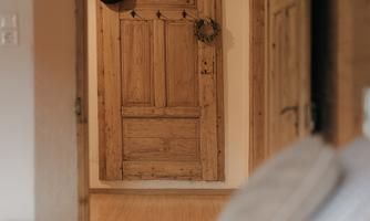 A rustic wooden door leads into a bright hallway. In the foreground, a bed and pillows are visible.