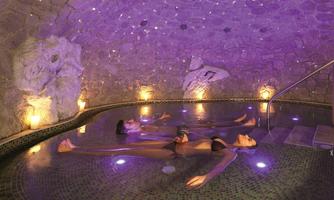 A relaxing spa environment with two people floating in the water. Soft, purple light and stone walls create a tranquil atmosphere.