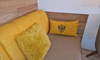 A cozy yellow sofa with a fluffy cushion and a decorative pillow. The wall is made of wood and creates a warm atmosphere.