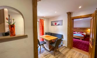 A cozy apartment with a dining area and a bedroom in the background. The decor is modern and friendly, with warm colors and plenty of wood.