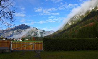 A picturesque mountain landscape with fog between the mountains and clear blue sky. In the foreground, there is a well-maintained garden with a hedge and a gate.