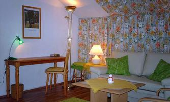 A cozy living space with a beige couch and a small wooden table. The walls are decorated with floral fabric, and there is a lamp as well as a desk.