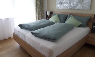 A modern bedroom with a large bed and bright colors. The bedding is in soft shades of green, and plenty of daylight streams through the window.
