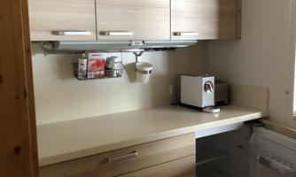 A modern kitchen with wooden-colored furniture and a clear, minimalist design. The equipment includes a refrigerator, a toaster, and plenty of storage space.