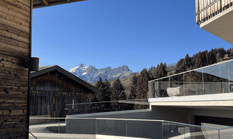 A modern balcony with a view of majestic mountains and a clear blue sky. Surrounded by forests and traditional wooden buildings.