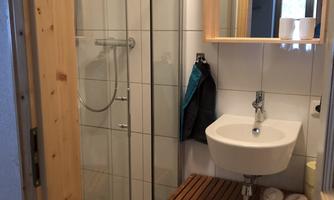 A small bathroom with a shower and a sink. There are wooden shelves for towels and toiletries.