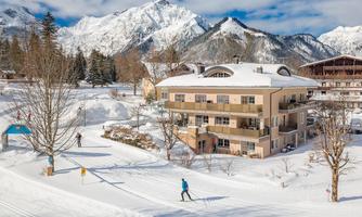A winter landscape with snow-covered mountains and a cozy building. A skier is going along the groomed trail.