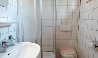 A small bathroom with tiles. On the left side: a wash basin and a shower cabinet. On the right side: the toilet. Very high above the toilet there are small windows.