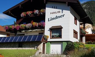 A charming house named "Haus Lindner" with colorful flowers on the balconies. In the foreground, there is a well-maintained garden with solar panels.