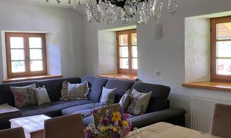 A cozy living space with a gray sofa and a large wooden dining table. An elegant chandelier hangs above the table, and a vase with colorful flowers is placed in the center.