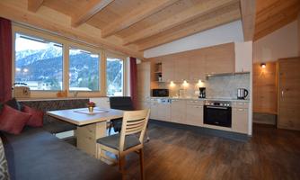 A modern kitchen unit with wooden paneling and large windows. The dining area is cozily furnished and offers a view of the mountains.