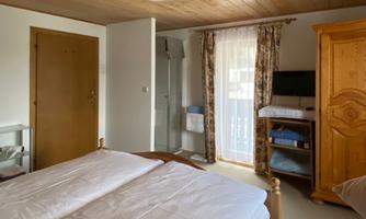 A cozy bedroom with two beds, a wooden wardrobe, and a television. The room has windows with curtains and access to a bathroom.