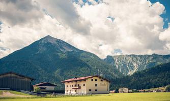A picturesque mountain panorama with tall mountains and a green meadow. In the foreground stand traditional rural buildings.