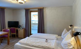 A modern room with two beds, a table, and a television. The window offers a view outside and there are bright curtains.