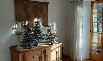 A cozy living space with a wooden cabinet displaying small Christmas trees. Large windows let in plenty of daylight.