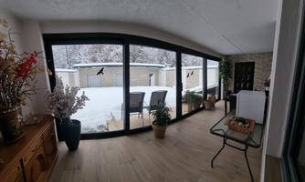 A bright conservatory with large windows overlooking fresh snow. The room features cozy chairs and plants.