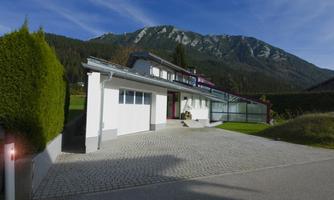 A modern house with large windows and a driveway. In the background, green mountains and a blue sky can be seen.