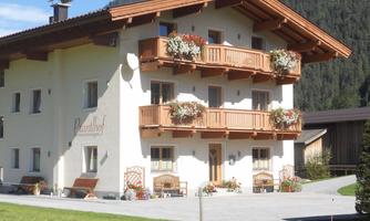 A cozy, two-story country house with wooden balconies and flower boxes. It is situated in a green environment with mountains in the background.