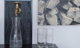 An elegant carafe of water sits on a table next to two glasses. In the background, a picture of stacked firewood hangs.