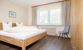 A modern bedroom with a wooden bed and large windows. The room is bright and welcoming, with a chair in the corner.
