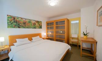 A bright bedroom with a large double bed and modern furniture. A colorful painting hangs on the wall, and a table with a plant stands in the room.